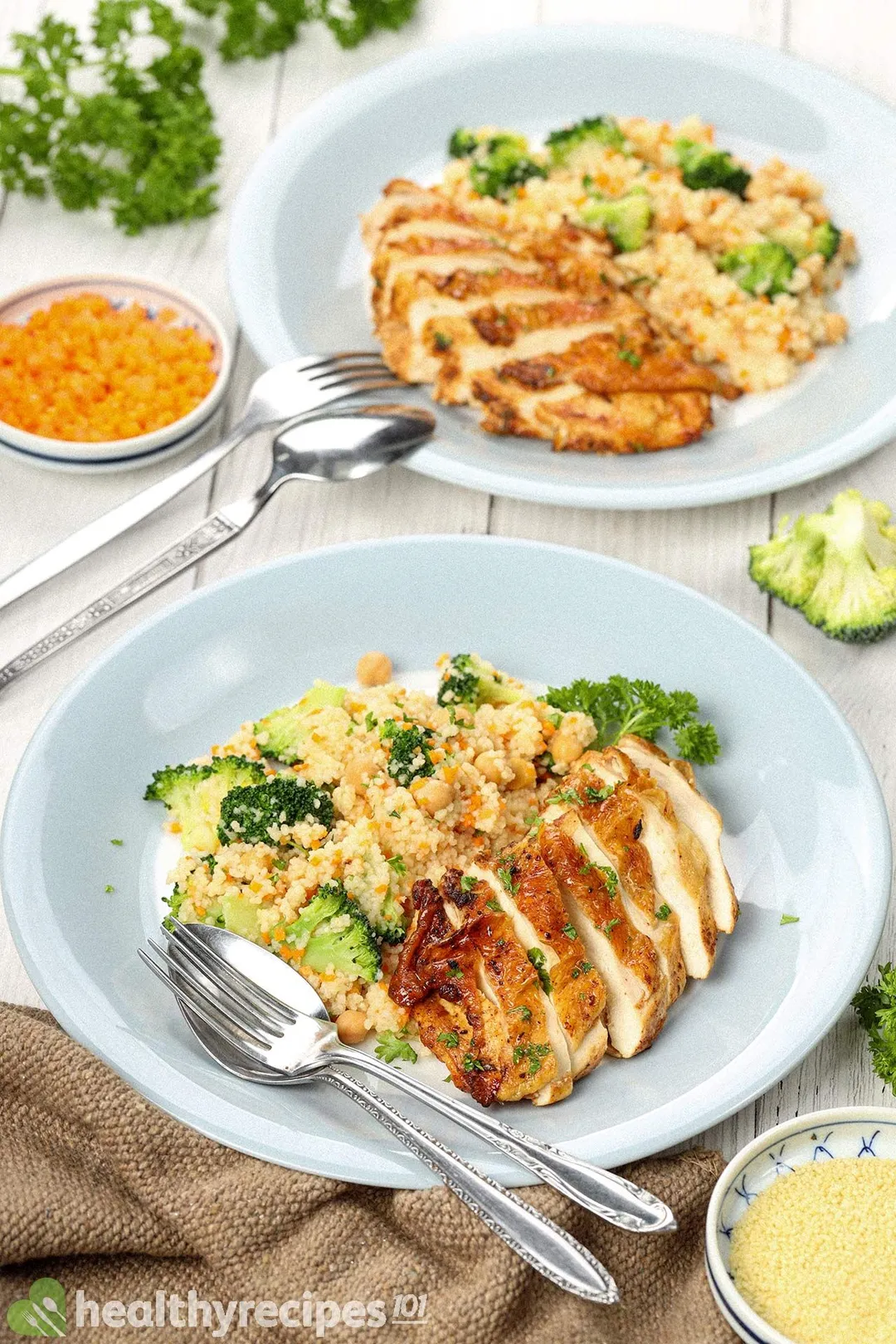 Two plates of chicken thighs and couscous laid near a brown tablecloth, a small bowl of uncooked couscous, a fork, and a spoon