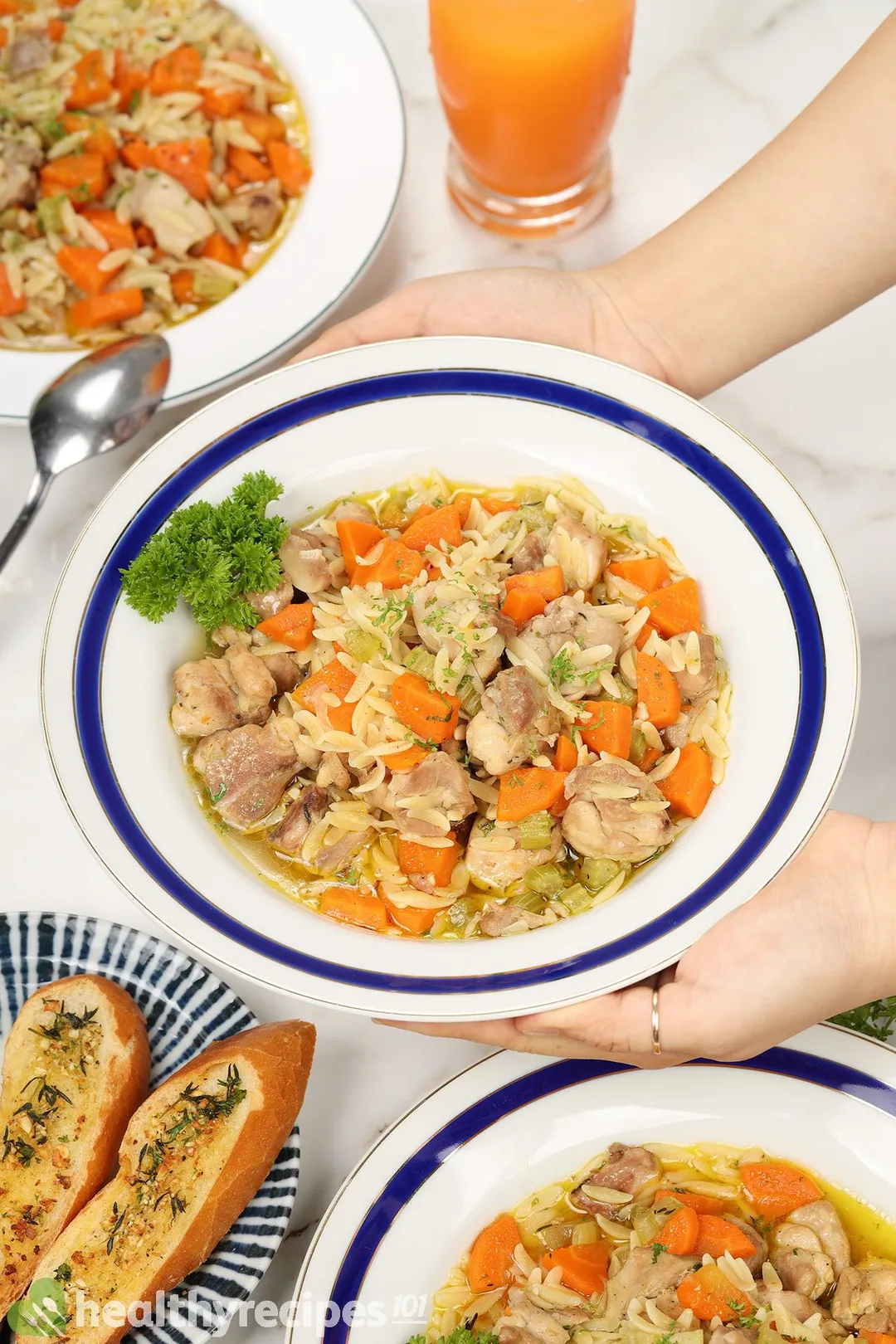 two hand holds a plate of cooked chicken orzo soup next to a glass of orange juice and a plate of garlic breads