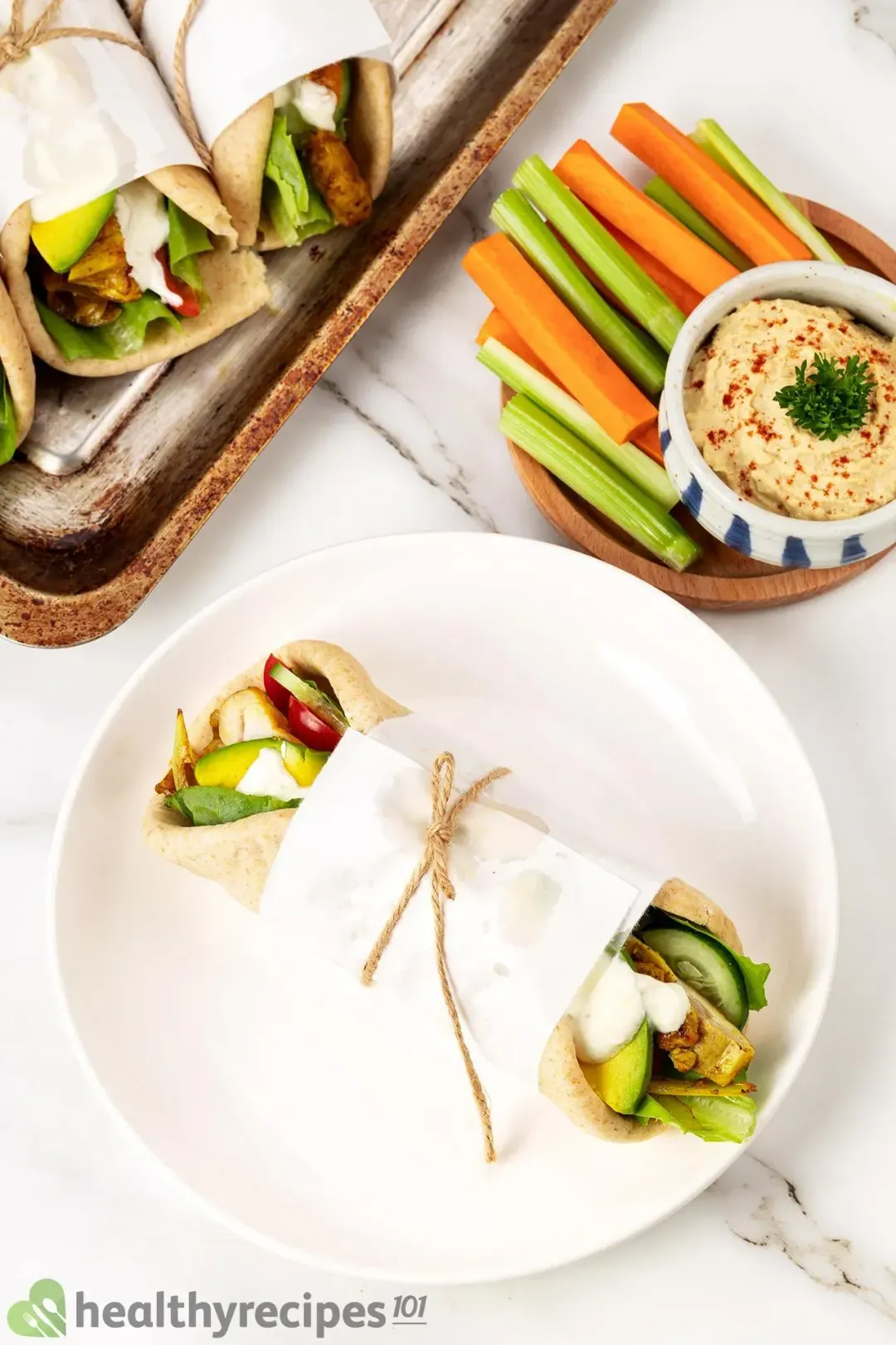 A wrapped-up chicken shawarma pita in a white plate, next to veg sticks and hummus and a baking tray