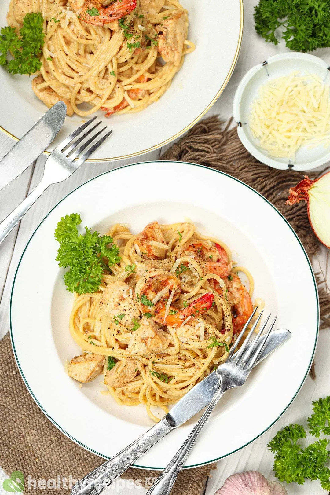 Two plates of chicken and shrimp alfredo with a metal knife and fork on the side, surrounded by a bowl of shredded cheese, parsley sprigs, a garlic bulb, and a halved onion