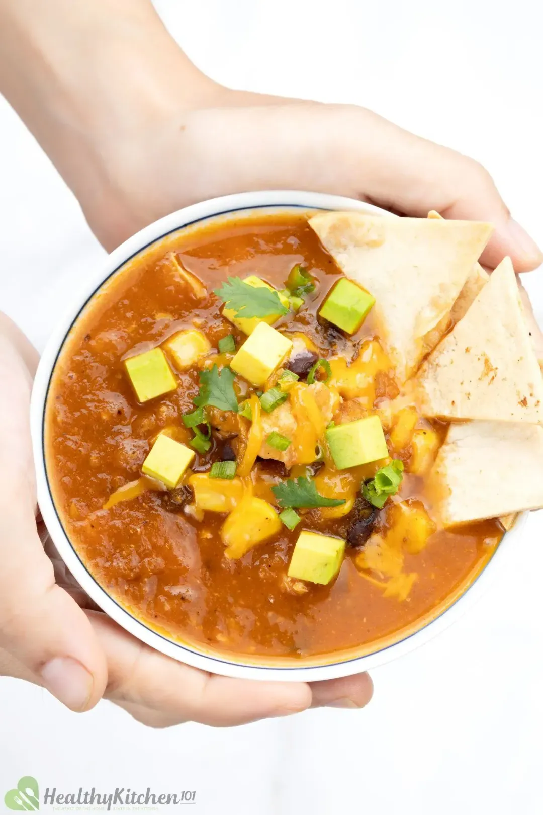 Two hands holding a bowl of chicken tortilla soup topped with zucchini cubes, shredded cheese and tortilla chips