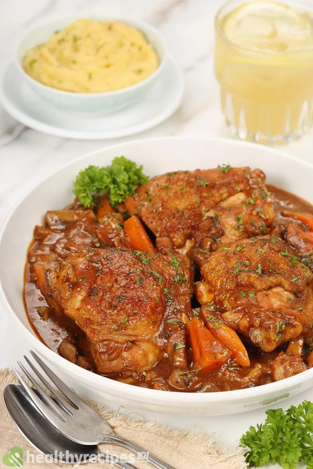 front shot of a bowl of cooked chicken thighs with a glass of lemonade and a bowl of mashed potato in background