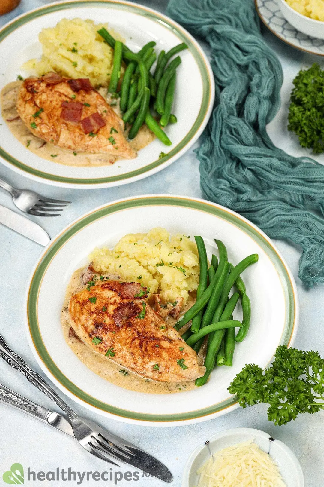 Two plates of cooked chicken breast, mashed potatoes, and green beans decorated by forks, dinner knives, and a peacock blue mesh cloth