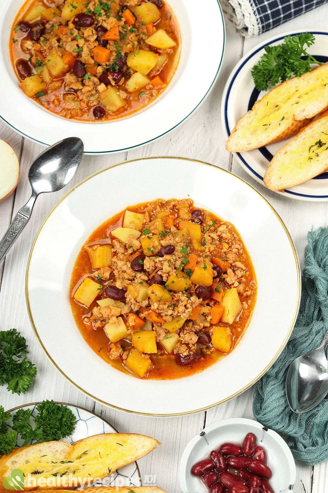 A high-angle shot of a deep white plate filled with chicken soup surrounded by a small dish of kidney beans, small slices of baguette, spoons, fresh parsley, and a blue mesh cloth