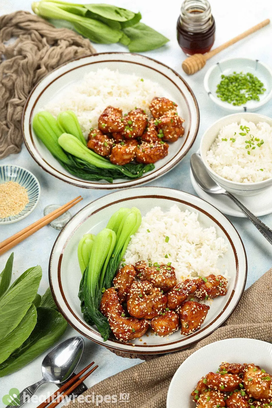 Two plates of honey chicken, white rice, and bok choy laid near a brown tablecloth, a spoon, a pot of honey, a bowl of white rice, and a pair of chopsticks