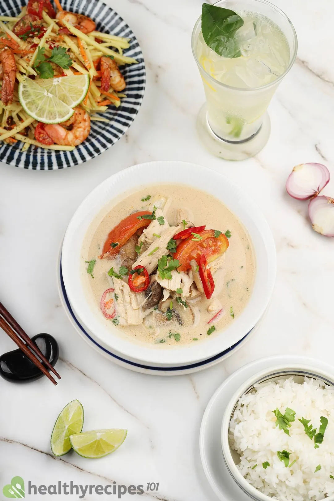 top view of a bowl of tom kha soup next to a glass of juice, a plate of mango salad and a bowl of cooked rice