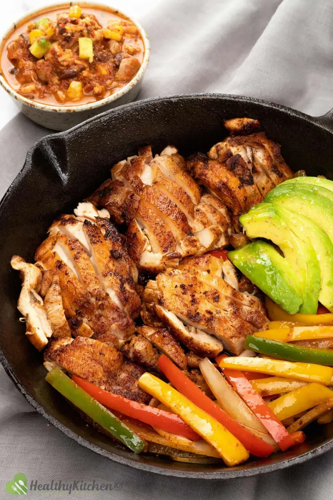 A skillet filled with pan seared chicken slices, bell pepper sticks, and avocado slices placed next to a bowl of soup