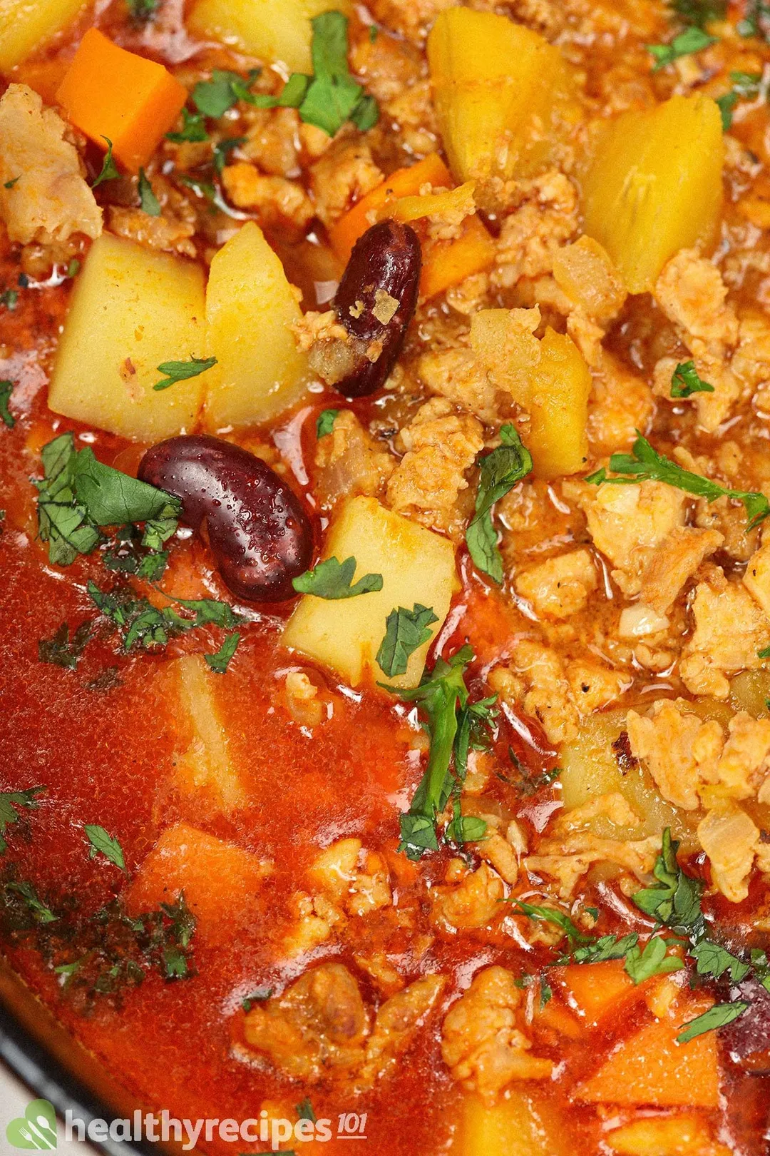 A close-up on a bowl of ground chicken soup, consisting of a dark red broth, coarsely diced carrots, cubed potatoes, ground chicken, and kidney beans