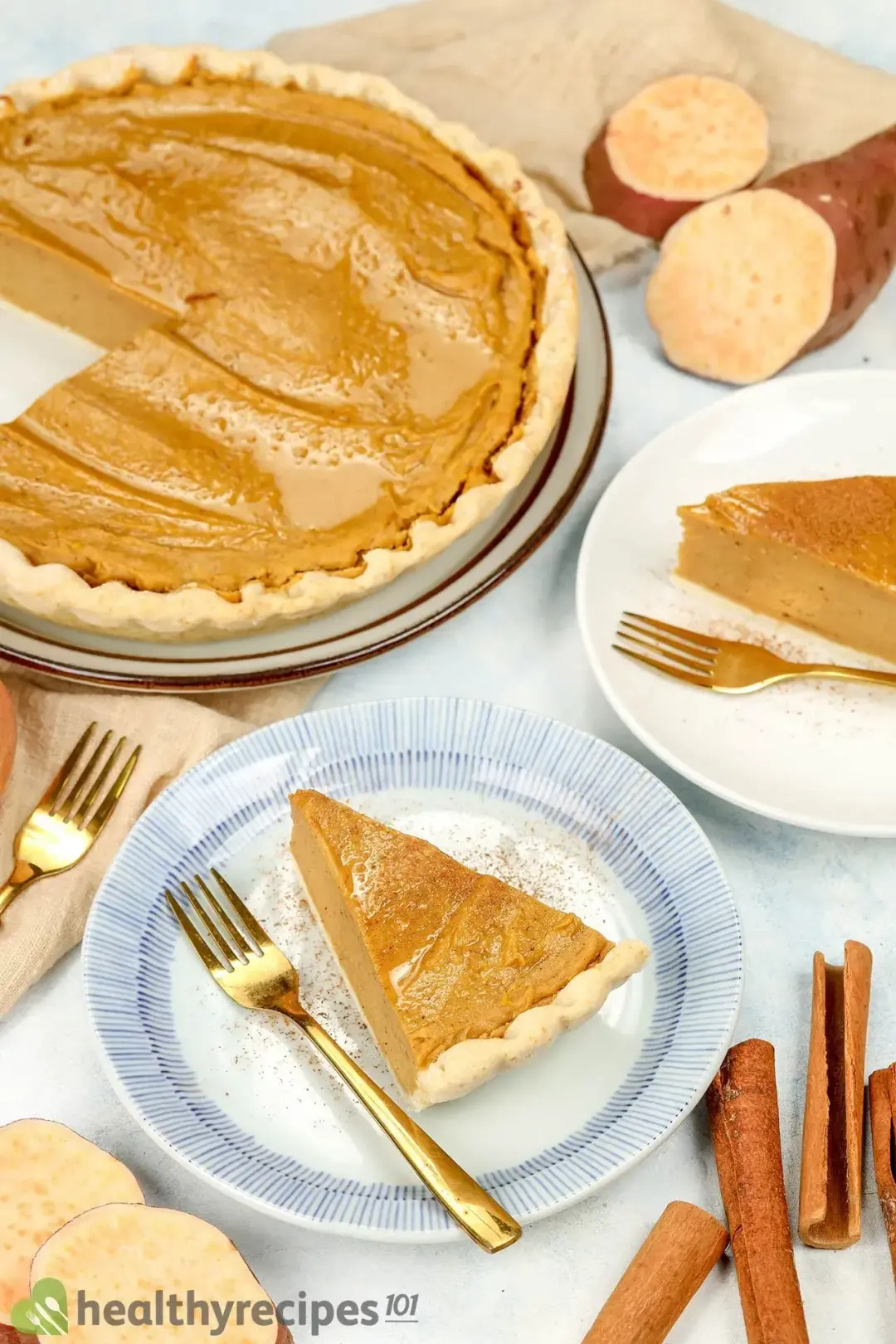 Slices of sweet potato pie and forks placed on plates near a large pie, surrounded by cinnamon sticks and sliced sweet potatoes