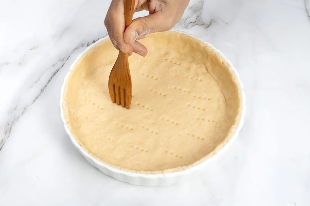 A hand using a wooden spoon to pierce airholes into an unbaked pie crust.