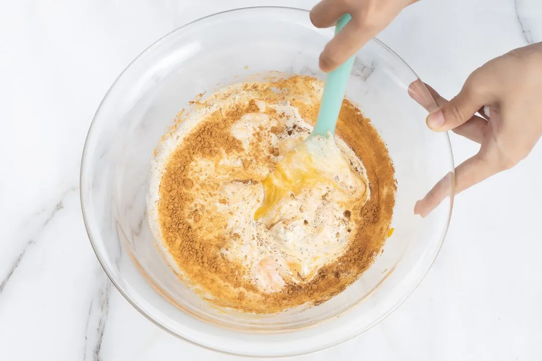 Two hands mixing a pumpkin mixture in a large glass bowl.