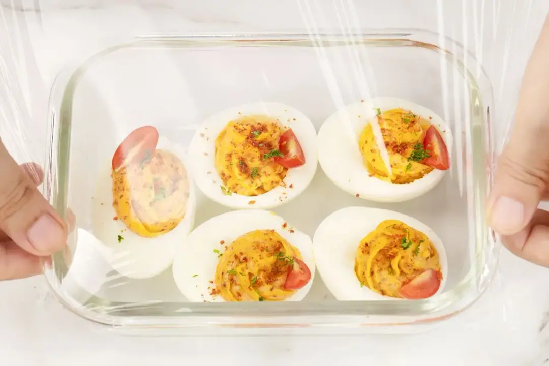Five deviled eggs in a clear container, with a clear cling wrap held over
