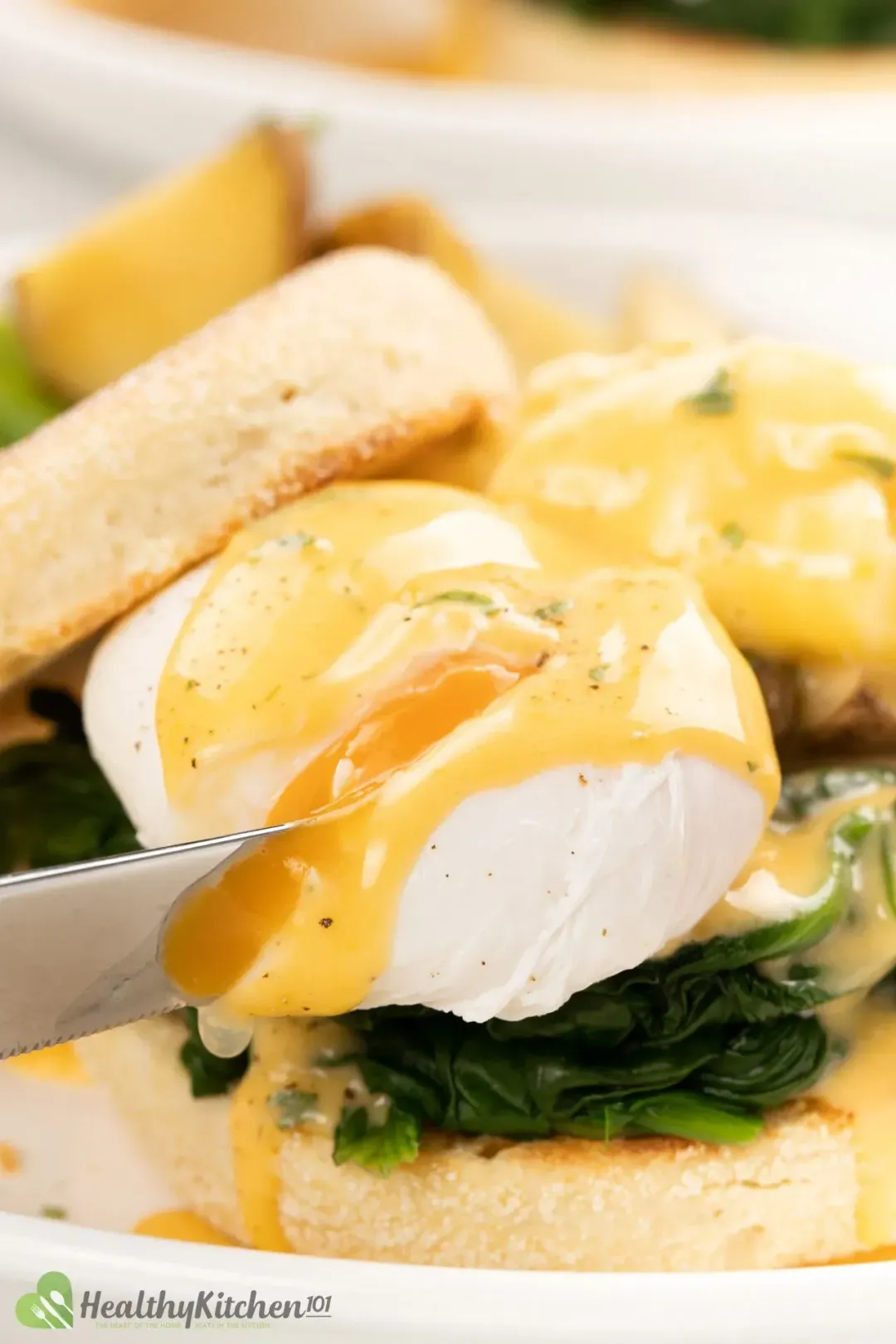 A knife slicing into a poached egg drenched in runny Hollandaise sauce over spinach and English muffin 