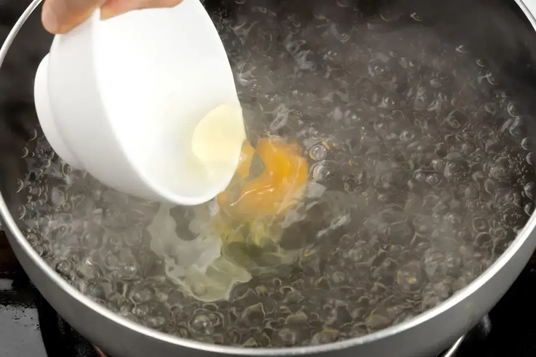 An egg being dropped from a white bowl into a pot of boiling water