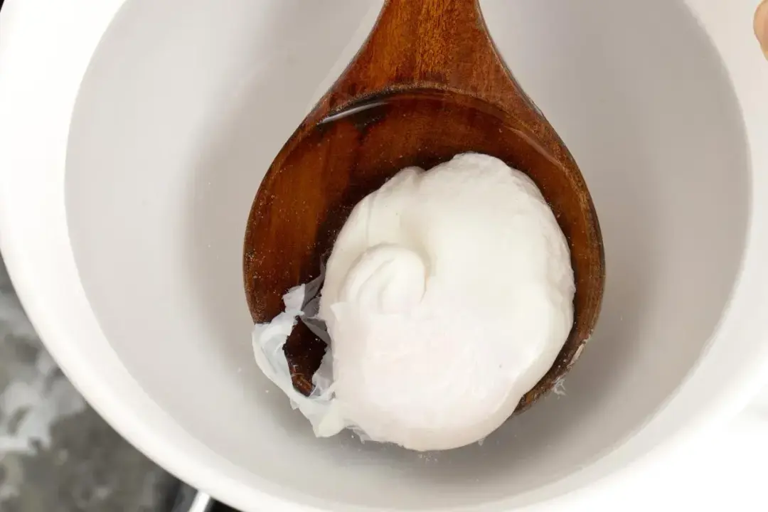 A wooden ladle holding a poached egg and dropping it into a white bowl of water