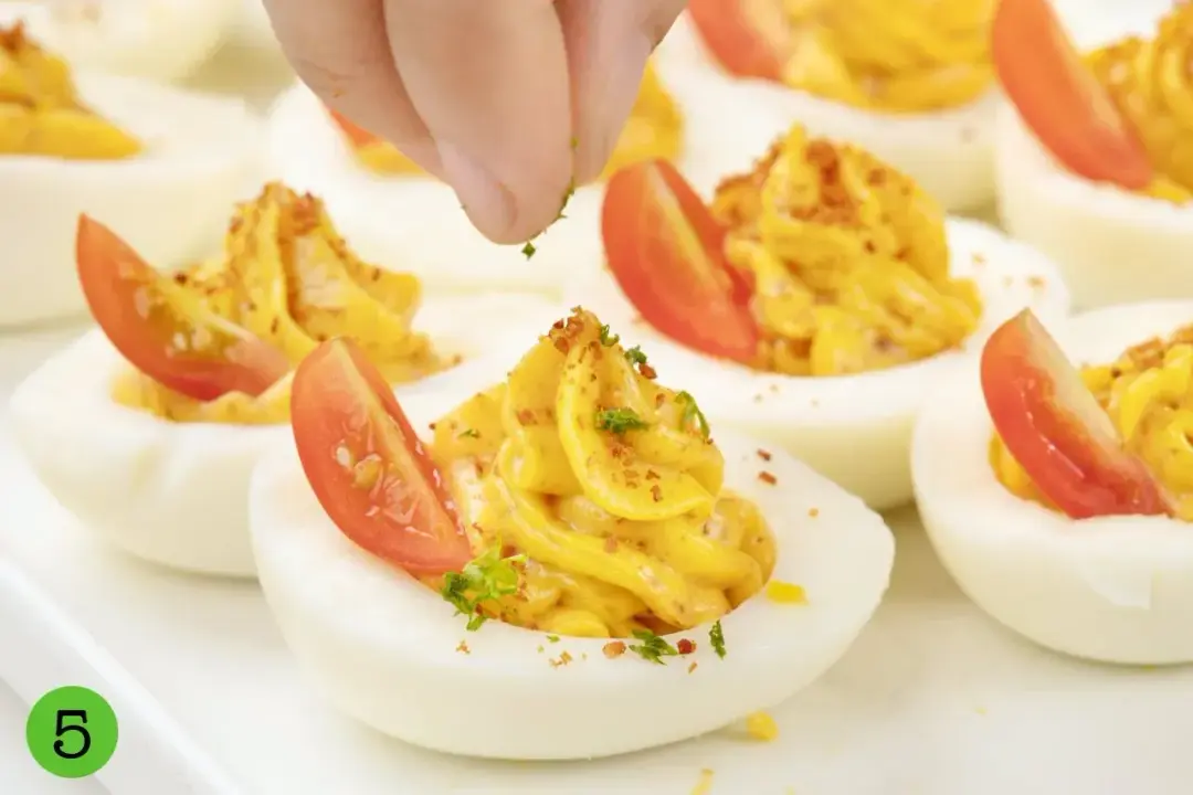 Deviled eggs with cherry tomatoes being topped with spices and herbs