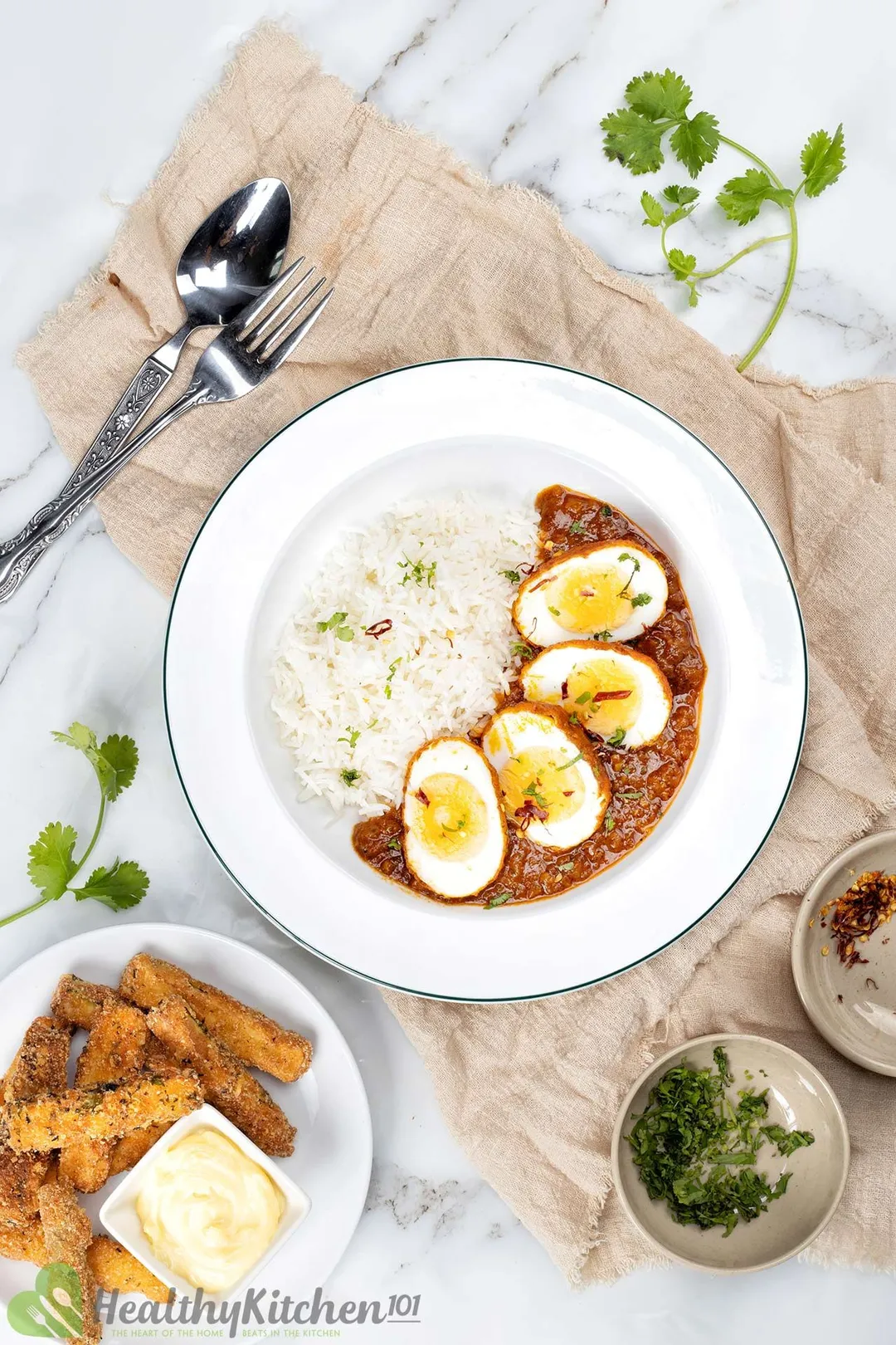 a plate of half eggs and rice next to a plate of fried zucchini, decorated with a fork and knife, two bowl of chopped cilantro, red pepper flakes