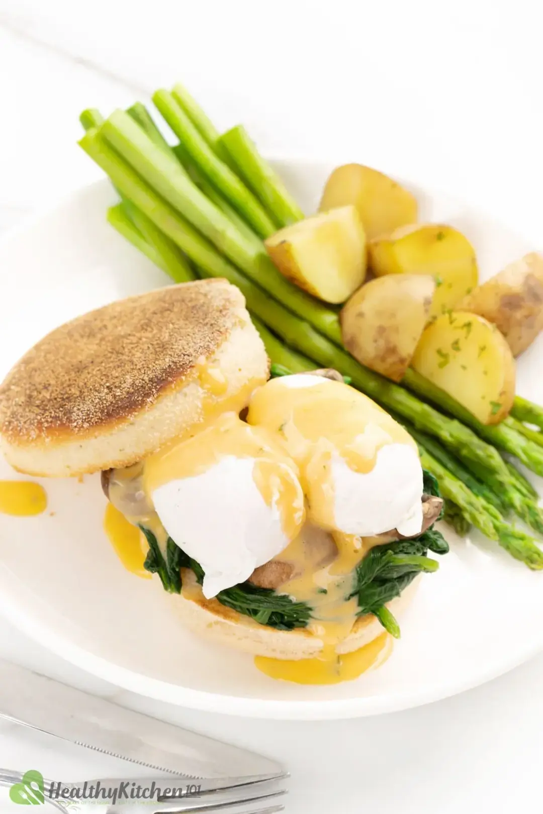 A white plate of English muffin holding poached eggs drizzled in Hollandaise and cooked spinach, served with potatoes and asparagus