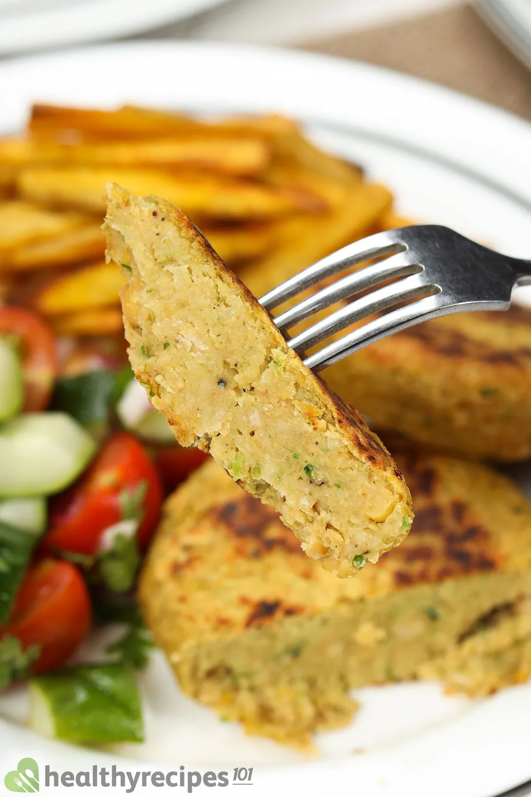 A fork piercing into a piece of chickpea patties with a plate of food in the blurred background.