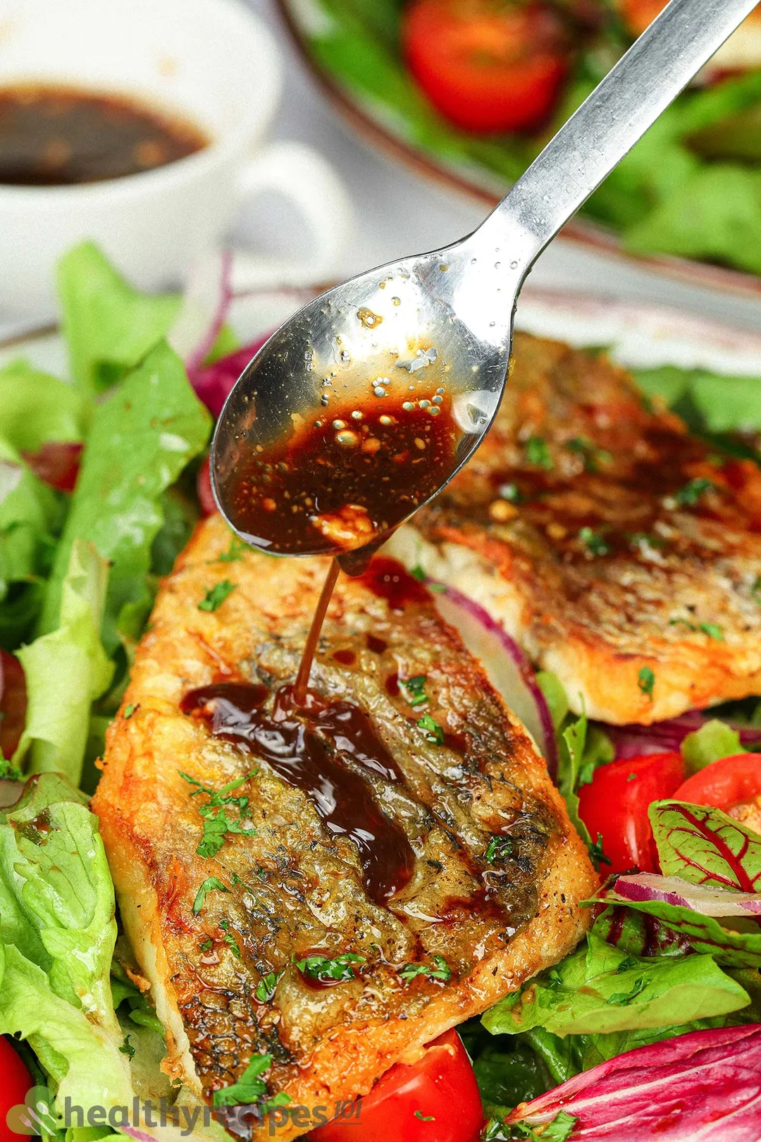 A spoon distributing balsamic vinegar onto pan-fried seabass fillets laid on a bed of lettuce.