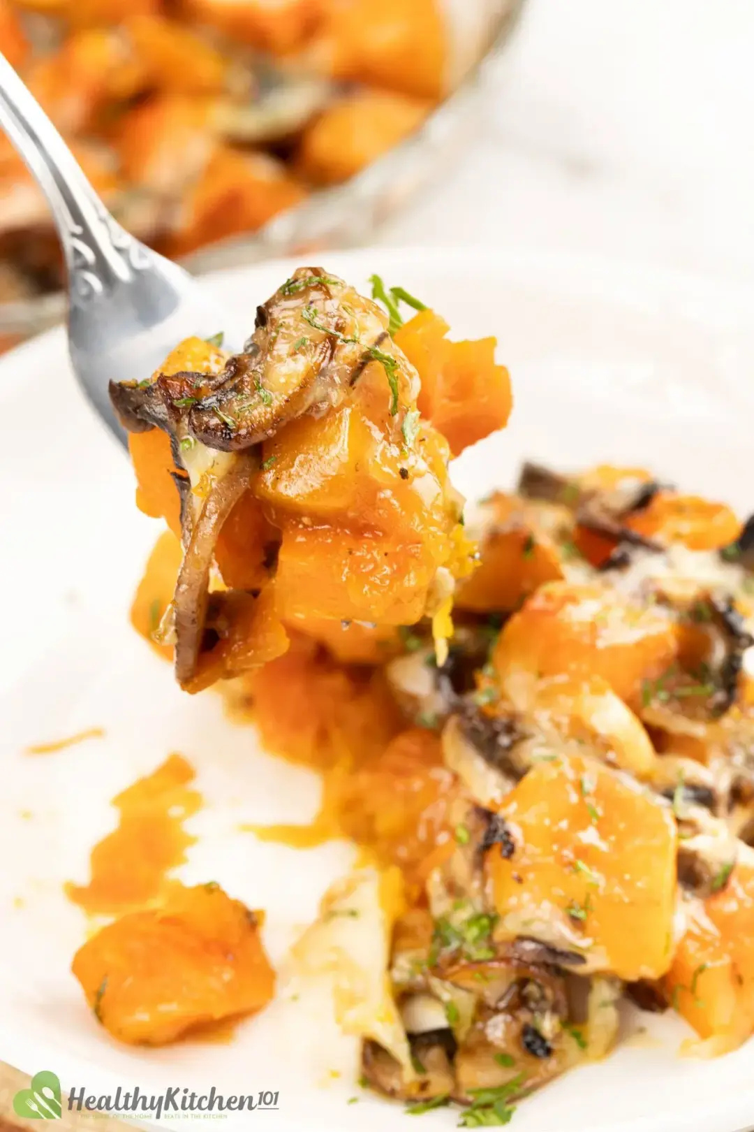  A fork holding pieces of roasted butternut squash and mushrooms, hovering over a dish of butternut squash casserole
