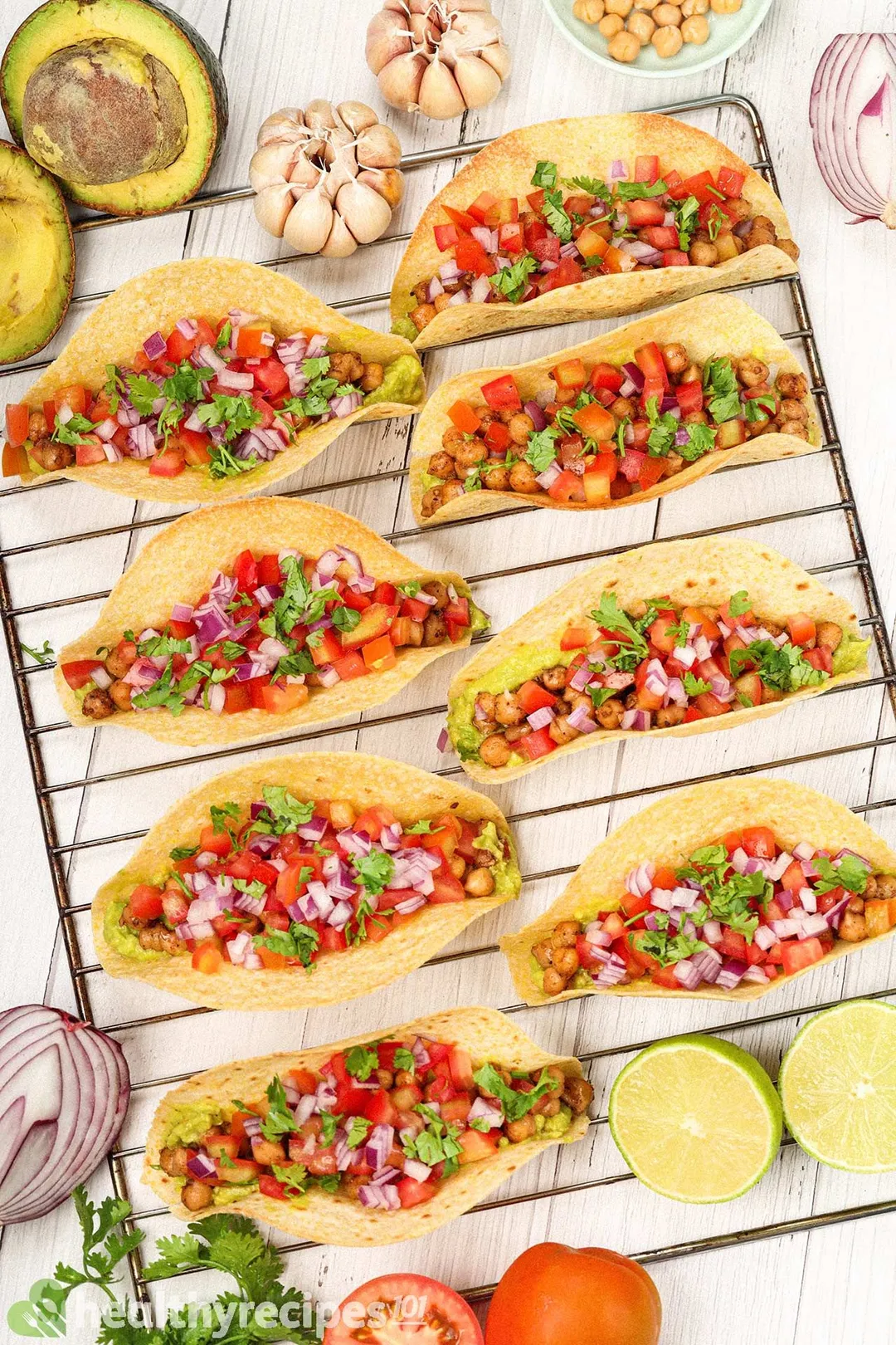 8 chickpea tacos laid on a rack near garlic cloves, red onion halves, avocado halves, and coriander.