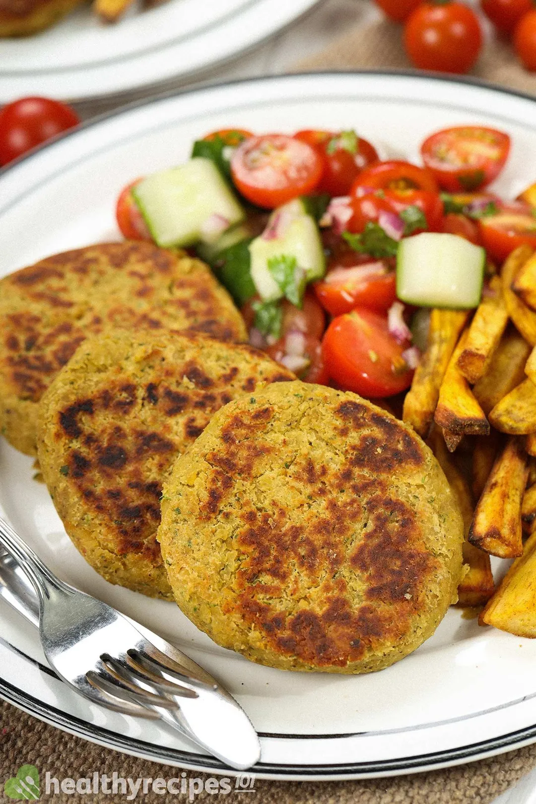 Three chickpea patties, sweet potato fries, cherry tomato halves, cucumber cubes, a fork, and a dinner knife laid on a white plate.and a 