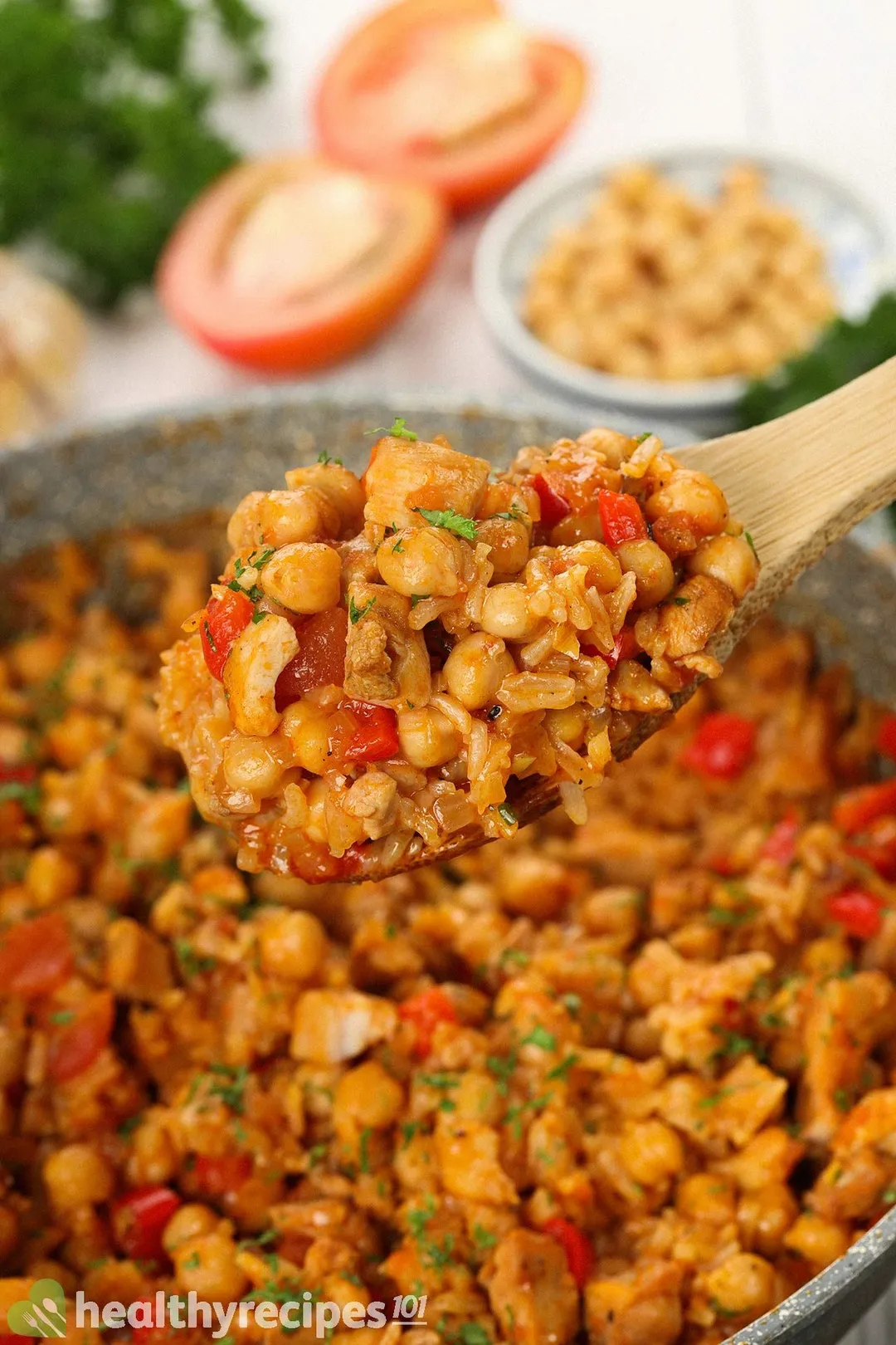 A wooden spatula scooping up chickpea, rice, diced bell pepper, and chopped cilantro with a large pan containing chickpea rice in the blurred background.