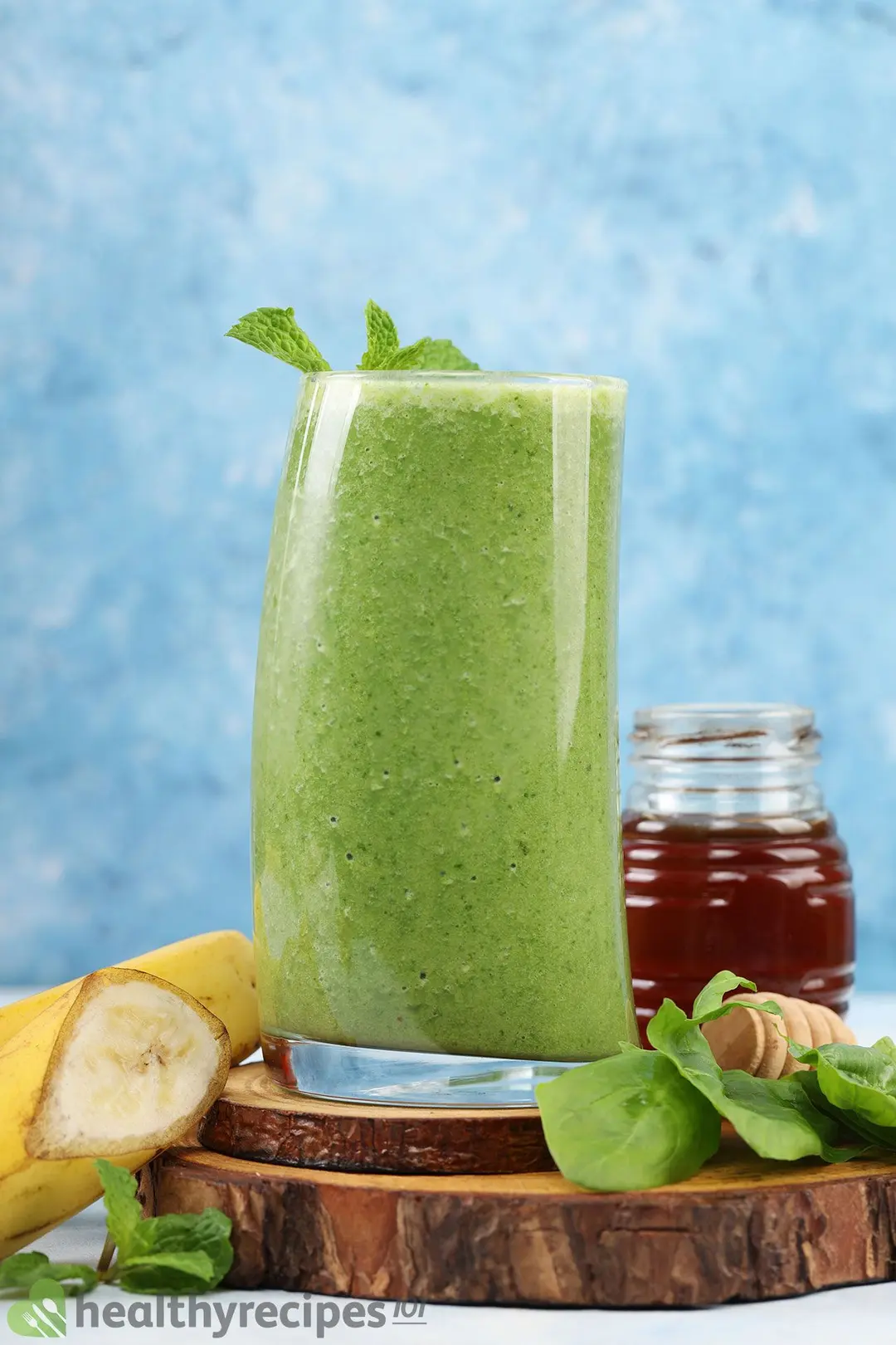 A tall glass of banana spinach smoothie placed on top of round wood cutting boards and near a sliced banana, fresh spinach, and a small honey pot