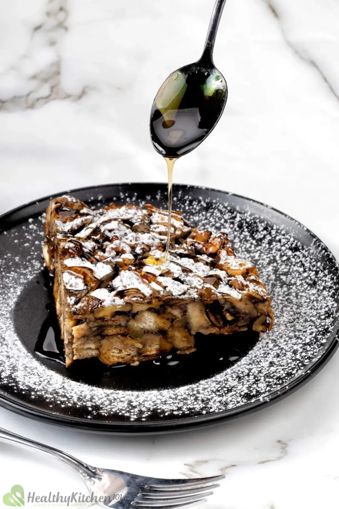 A black plate of French toast casserole dusted with powdered sugar, drizzled with maply syrup on top