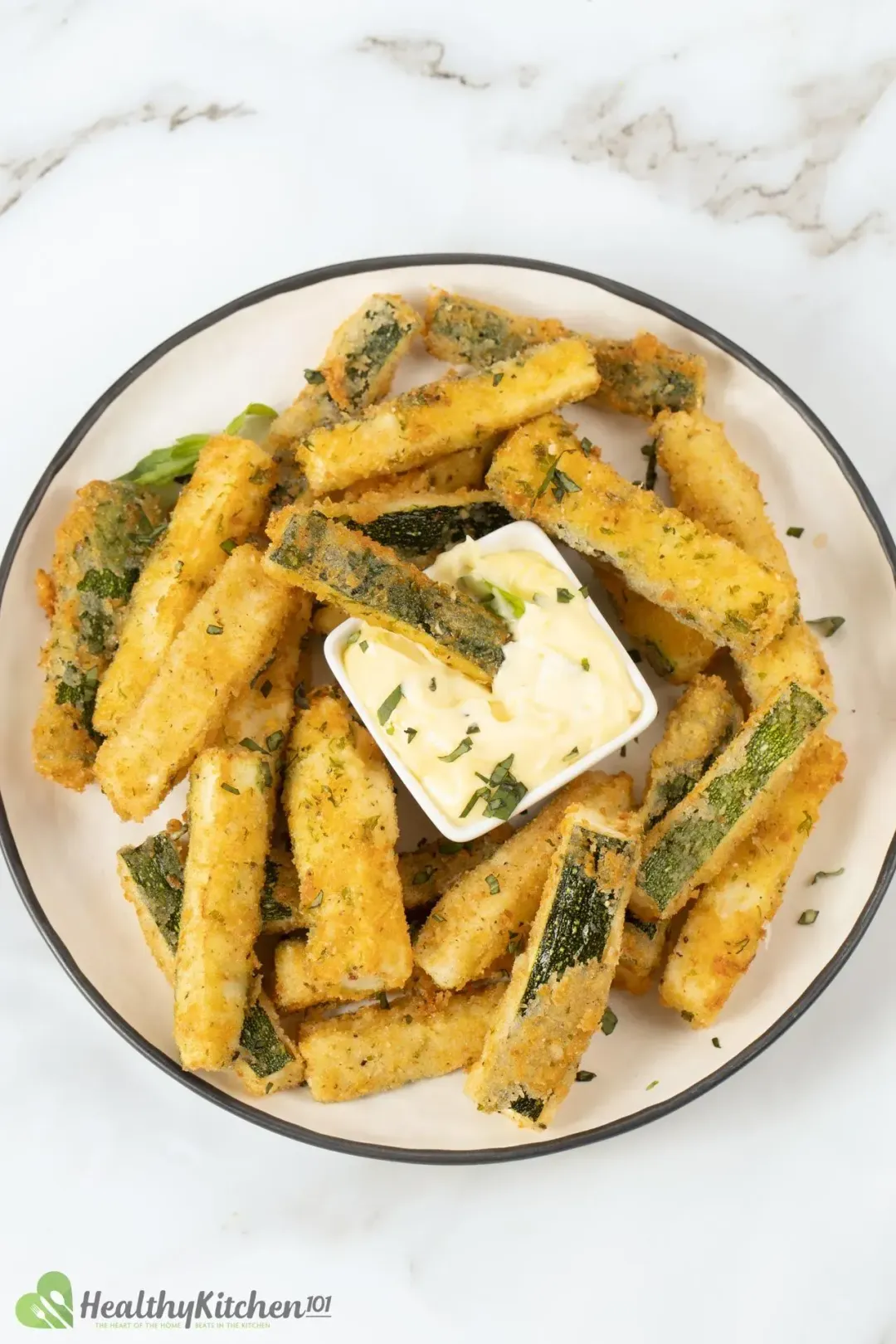 A plate full of deep-fried zucchini fries topped with herbs, surrounding a mayo dip