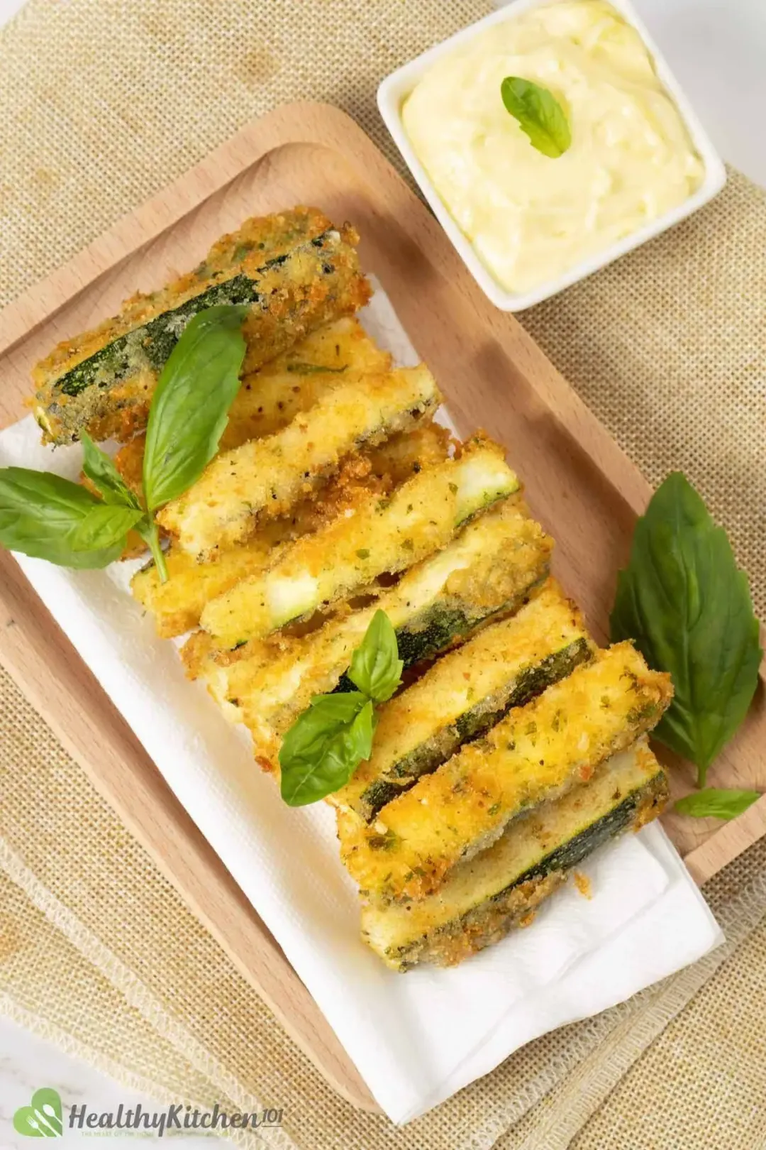 A plate of zucchini fries lined with white napkins, next to a dipping sauce garnished with herbs