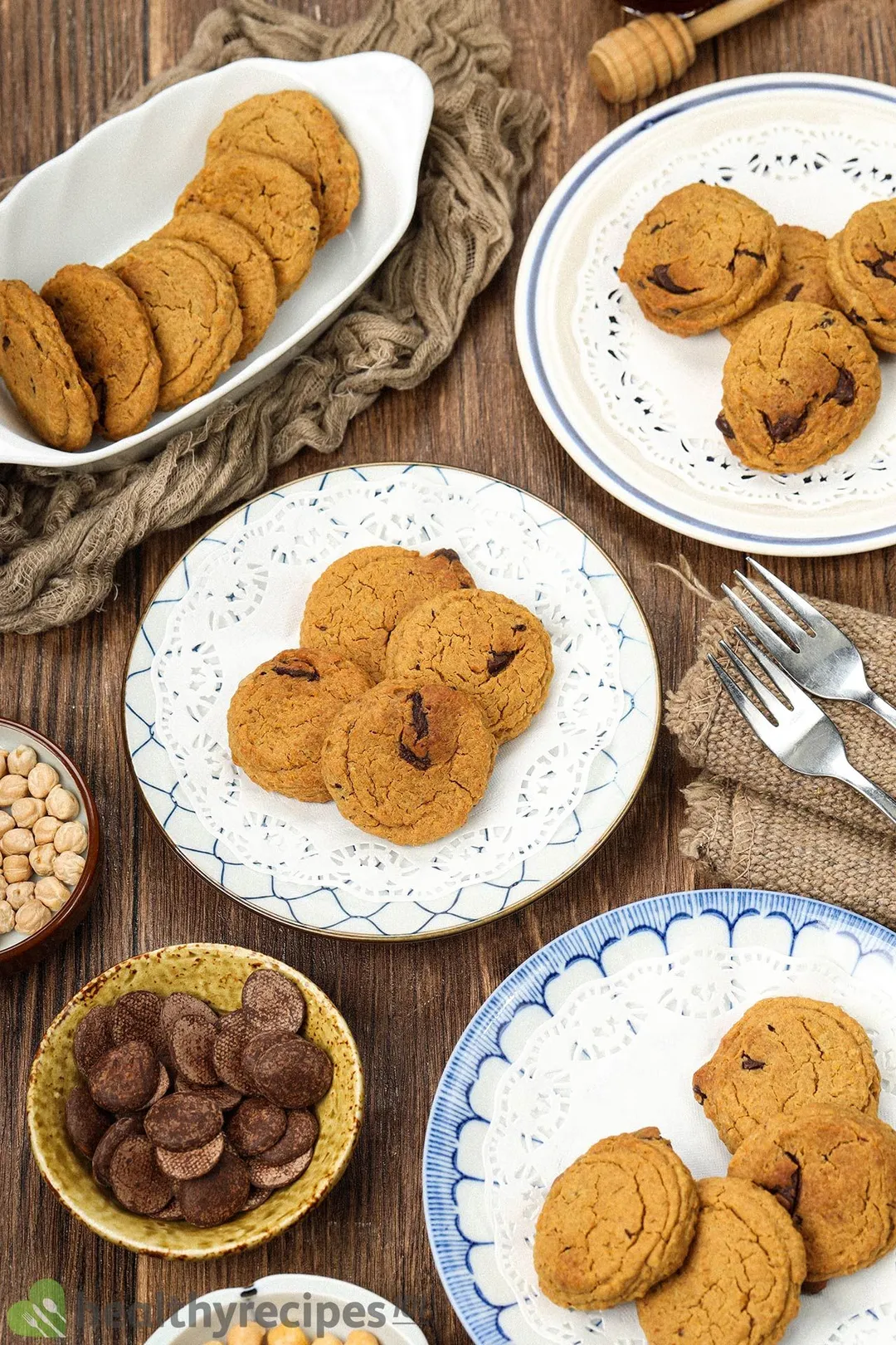 Plates of chickpea cookies laid on a wooden surface and surrounded by plates of chocolate chips, chickpeas, and forks.