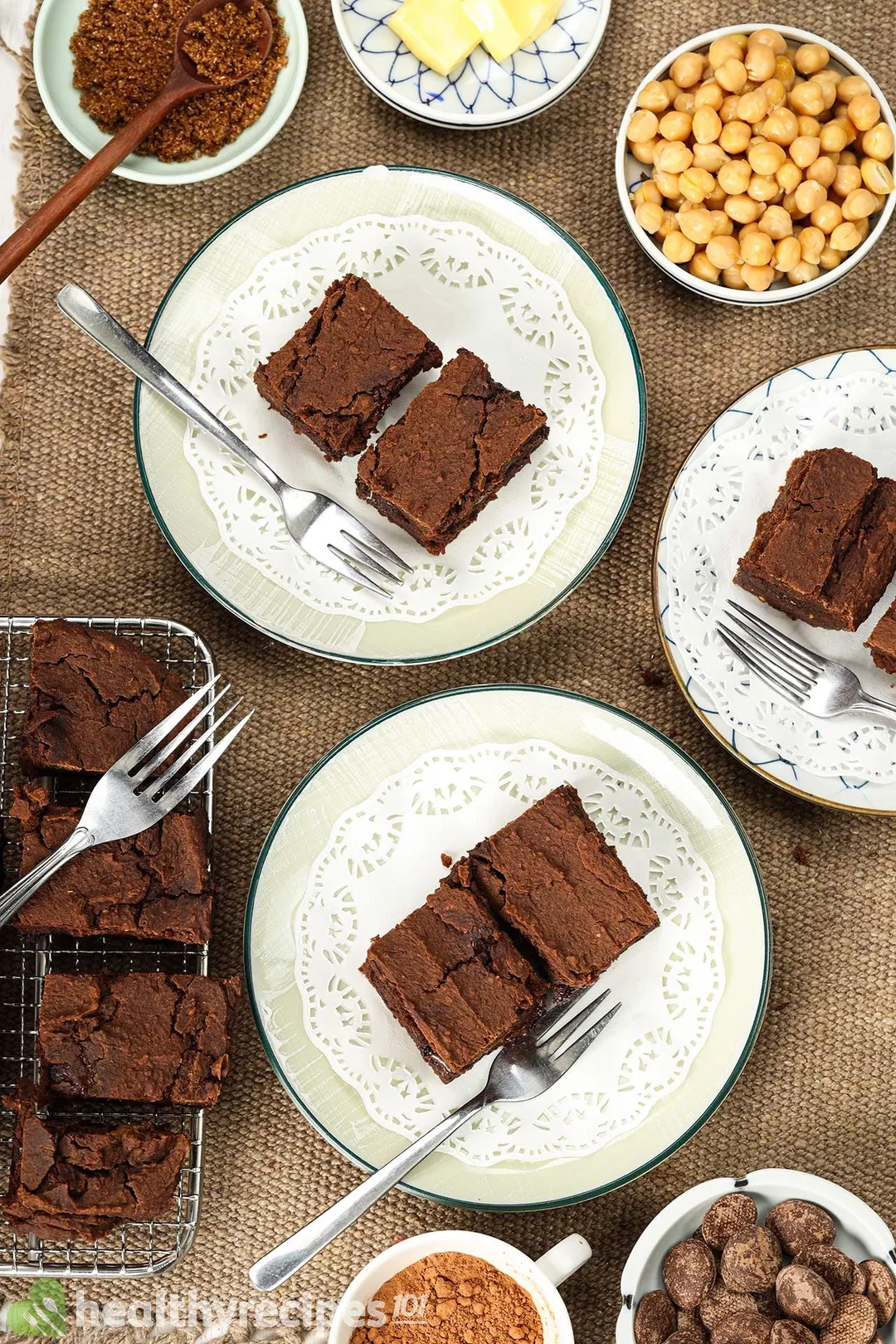 Plates of chickpea brownies with forks on the side laid near a small disk of chickpea, cocoa powder, chocolate chips, and butter cubes.