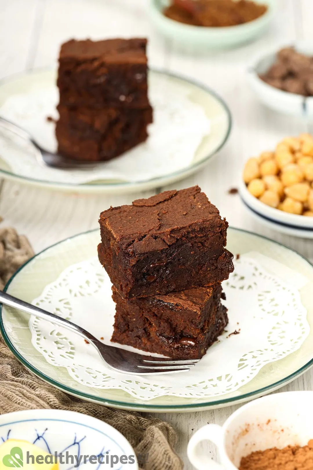 Two chickpea brownie squares stacked on top of each other with a fork laid on the side and a similar dish on the background.
