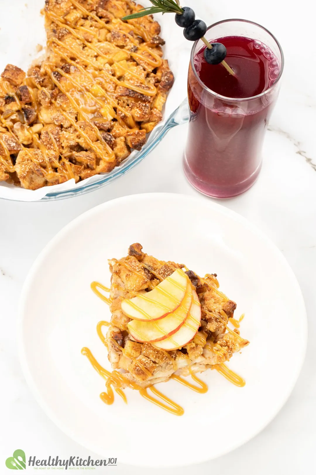 a white plate of bread pudding with sliced apple next to a glass of blue berry juice