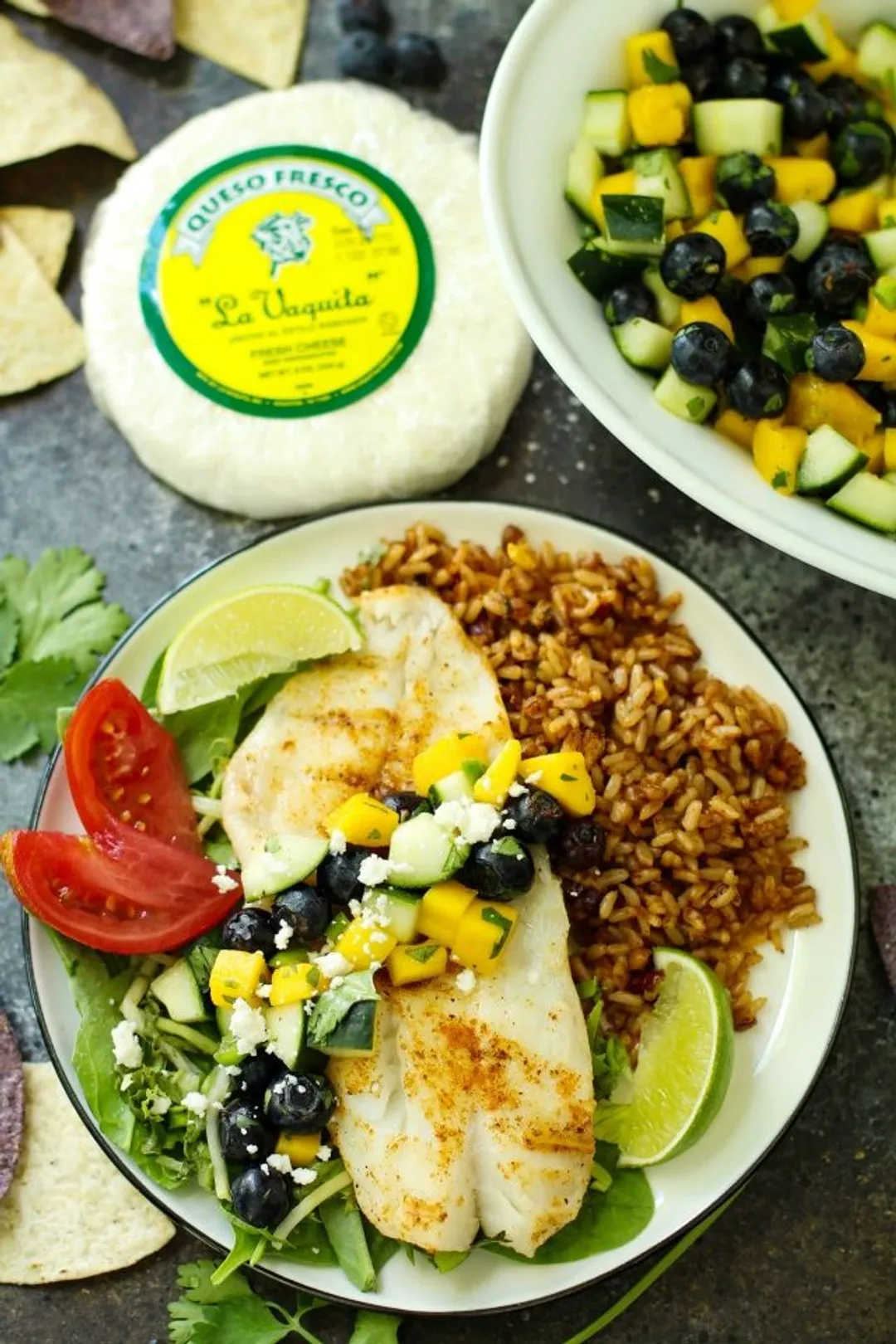 A plate of brown rice, cooked orange roughy, cilantro, lime wedges, tomato wedges, corn, and black beans laid near a block of La Vaquita Queso Fresco Cheese