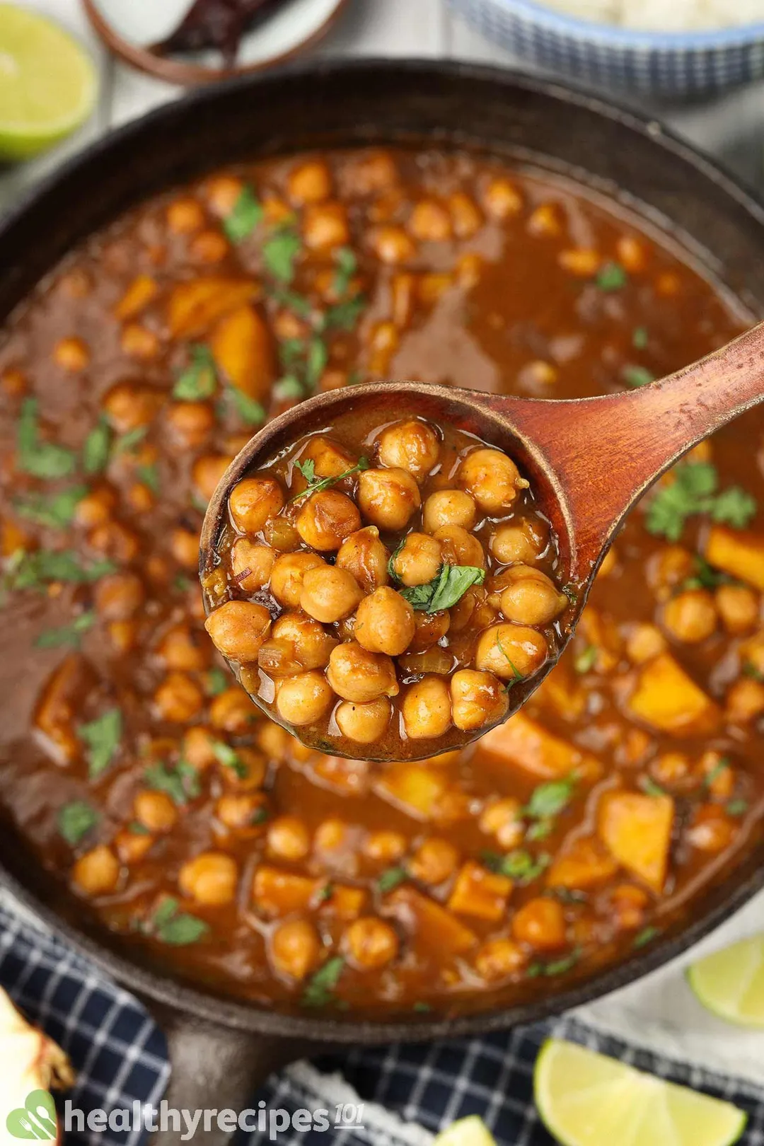 A wooden ladle scooping up chickpea curry with a skillet of chickpea curry in the background.