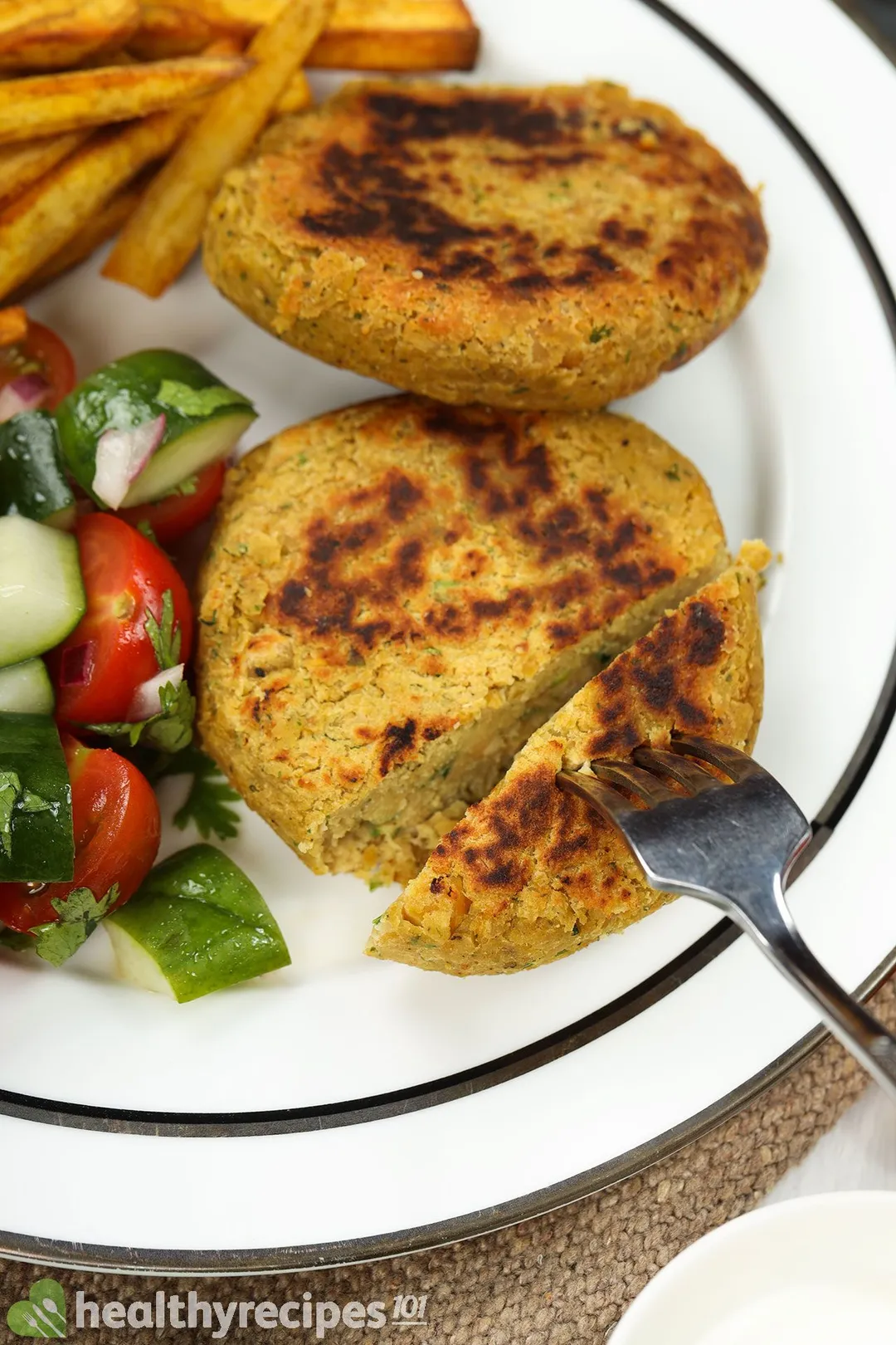 A fork piercing into a chickpea pattie laid on a white plate near another chickpea pattie, cherry tomatoes, and sweet potato sticks.