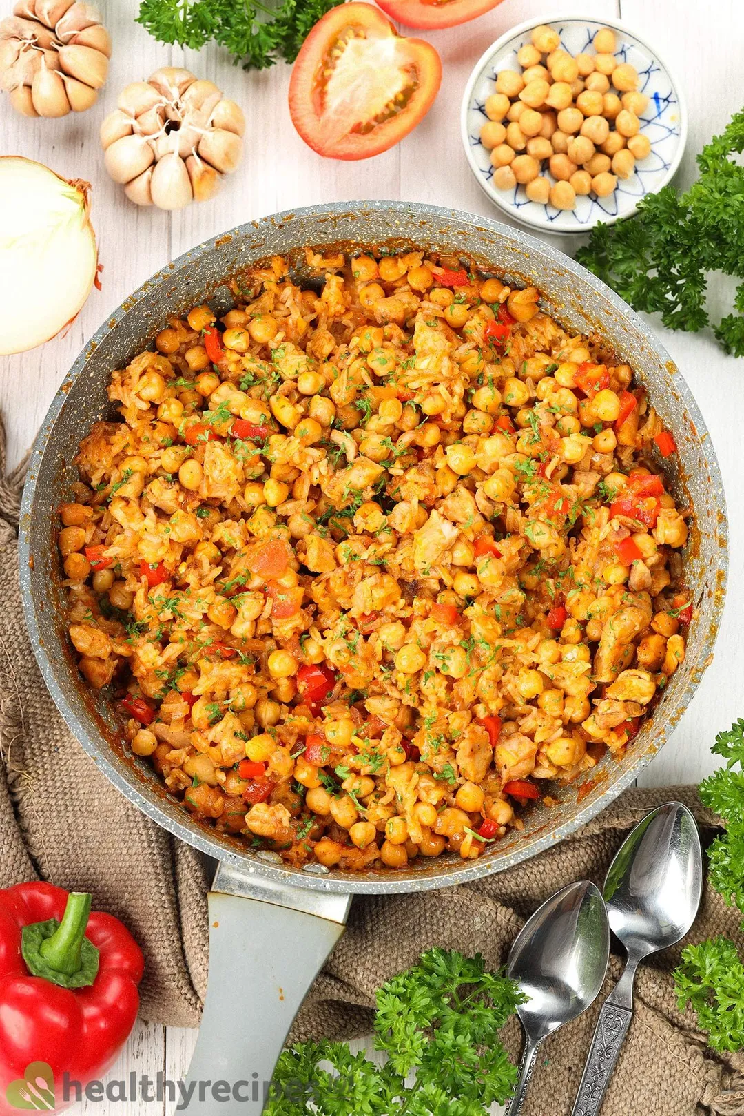 A large pan filled with chickpea, rice, diced bell pepper, and chopped herbs surrounded by garlic cloves, tomato halves, bell pepper, fresh parsley, spoons, and a brown cloth.