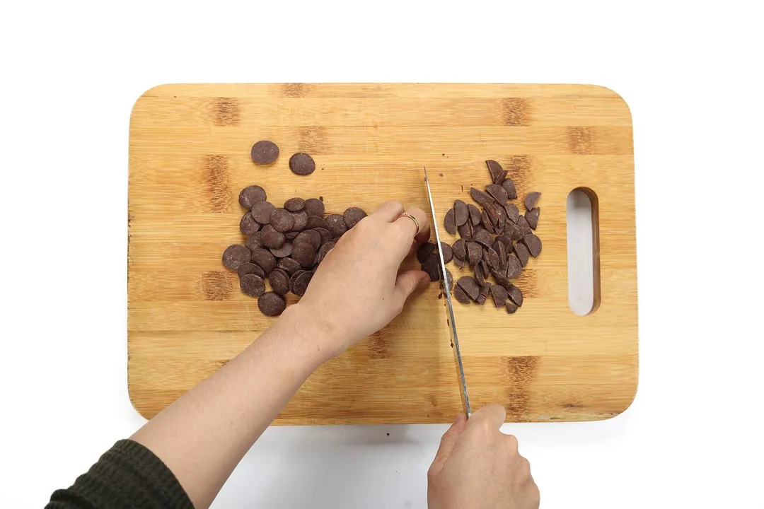 Two hands chopping chocolate chips into smaller pieces on a wooden chopping board.