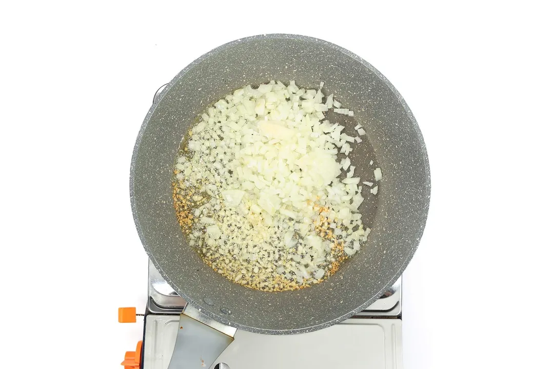 A large pan cooking diced onion and minced garlic on a portable gas stove.