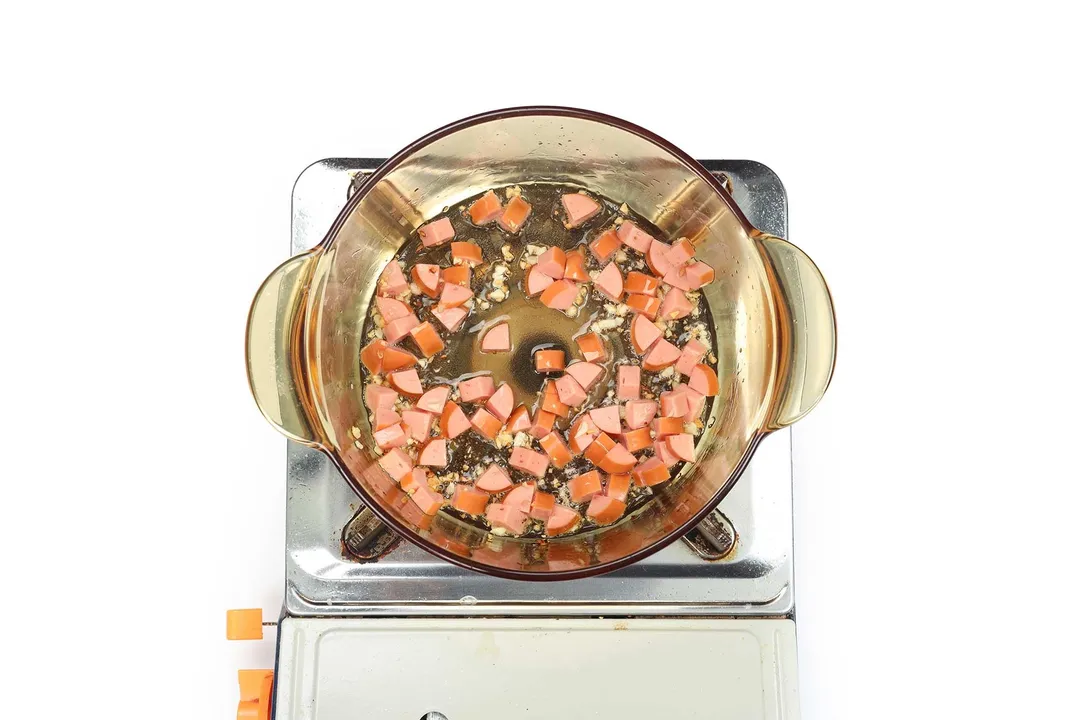 A clear, glass saucepan cooking diced sausage on a portable gas stove.