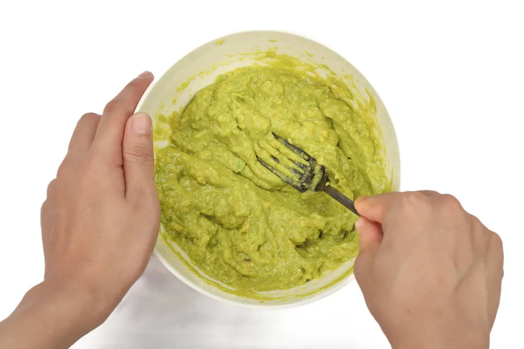 Two hands mashing avocado in a white bowl with a fork.