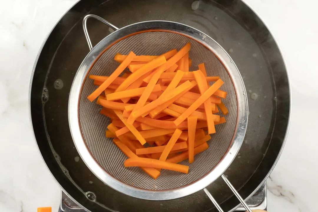A strainer picking up julienned carrots from a boiling pot of water.