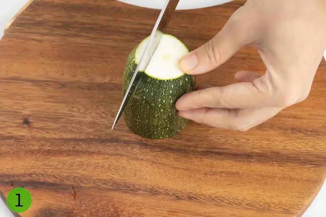 A zucchini cylinder stood straight on a wooden cutting board by a hand, with a knife blade placed on top