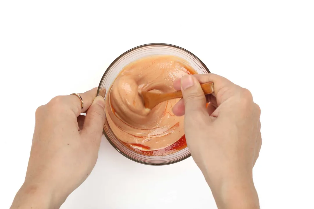 Two hands using a wooden spoon to mix a milky orange sauce in a small glass bowl.