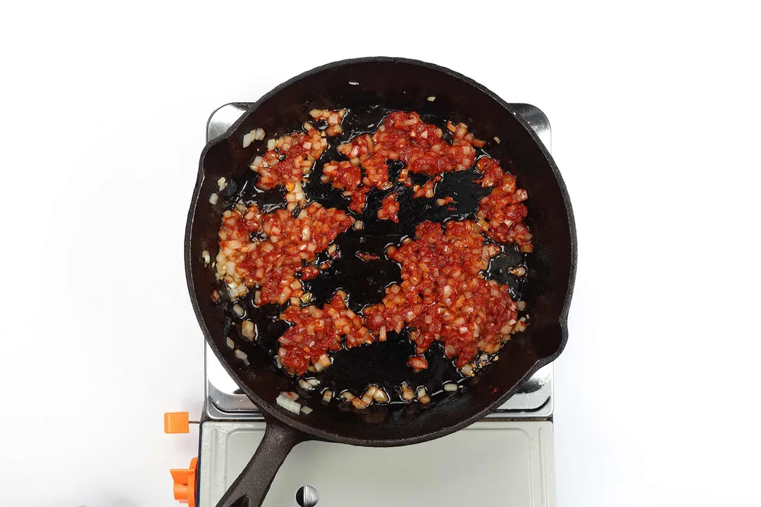 A skillet cooking diced onion coated with tomato paste on a portable gas stove.