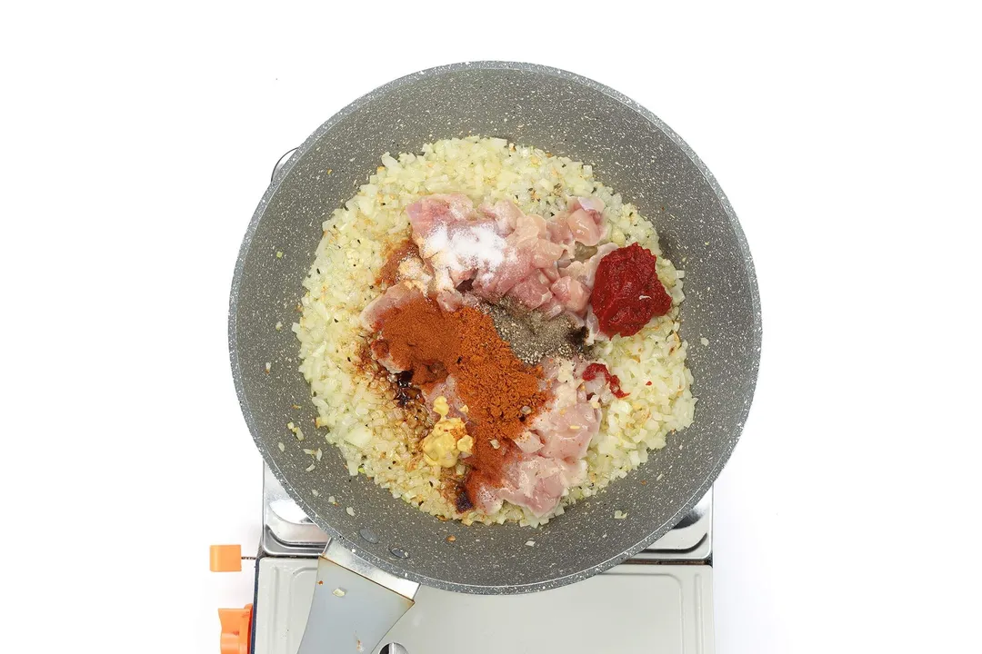 A pan cooking diced onion and cubed raw chicken with tomato paste, Cajun seasoning, and various other condiments.