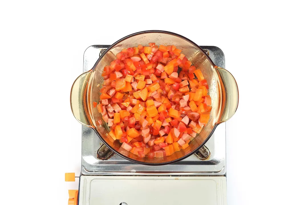 A glass saucepan cooking diced tomatoes, coarsley diced carrots, and diced sausage on a portable gas stove.